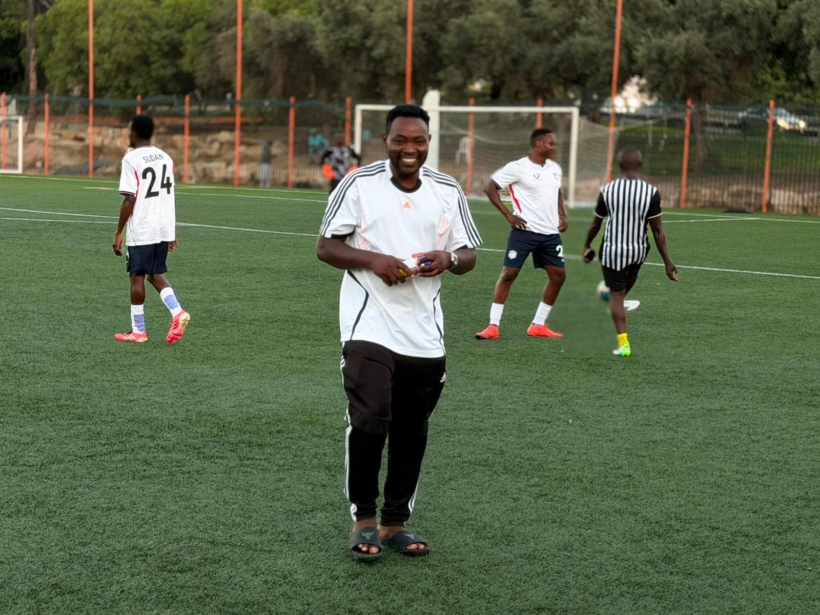A man smiling on the field as players warm up behind him.