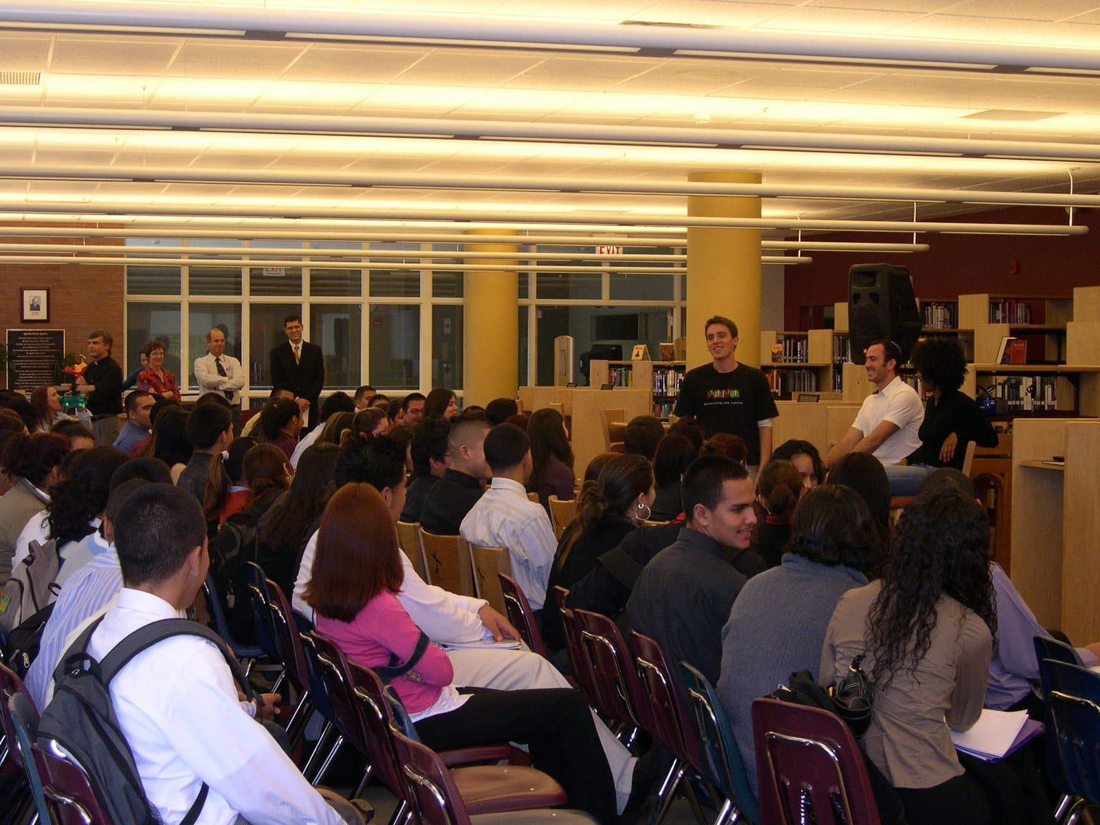 A delegation addressing a large seated student audience in a school library.