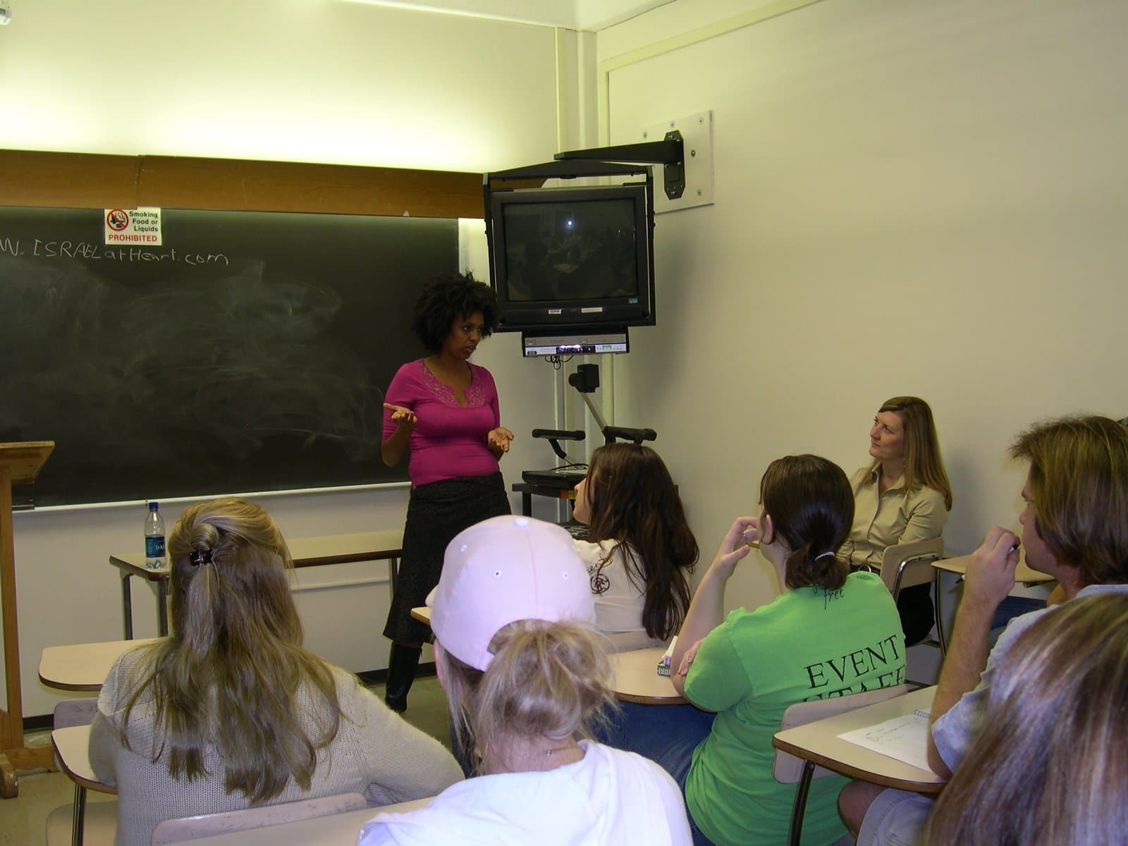 An Ethiopian-Israeli delegate speaking and gesturing to a seated class, a chalkboard reading israelatheart.com behind her.