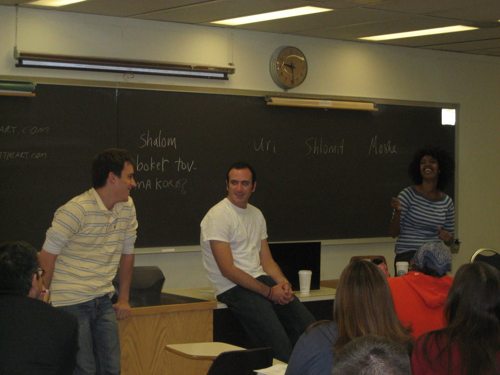 Two delegates seated on a desk beside a standing Ethiopian-Israeli delegate, their first names written on the chalkboard behind them.