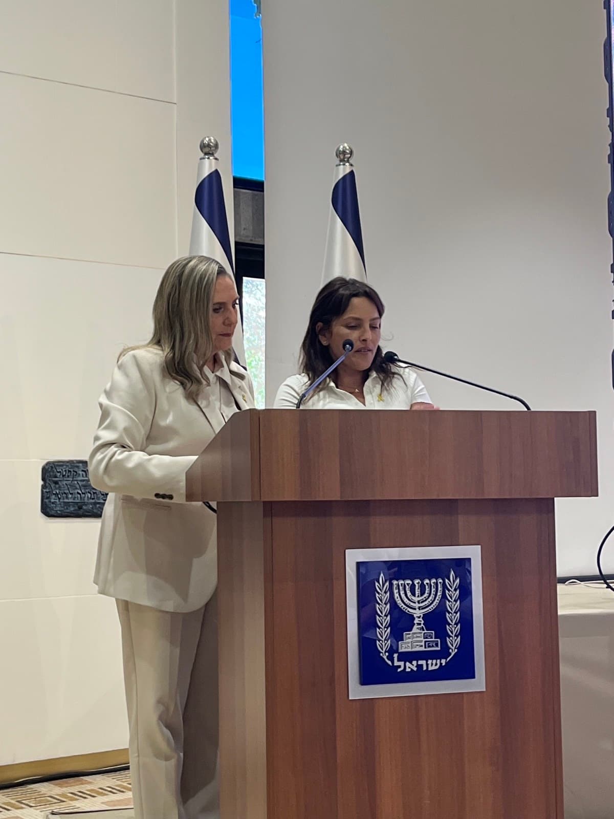 First Lady Michal Herzog and former hostage Amit Soussana at a podium bearing the emblem of the State of Israel.