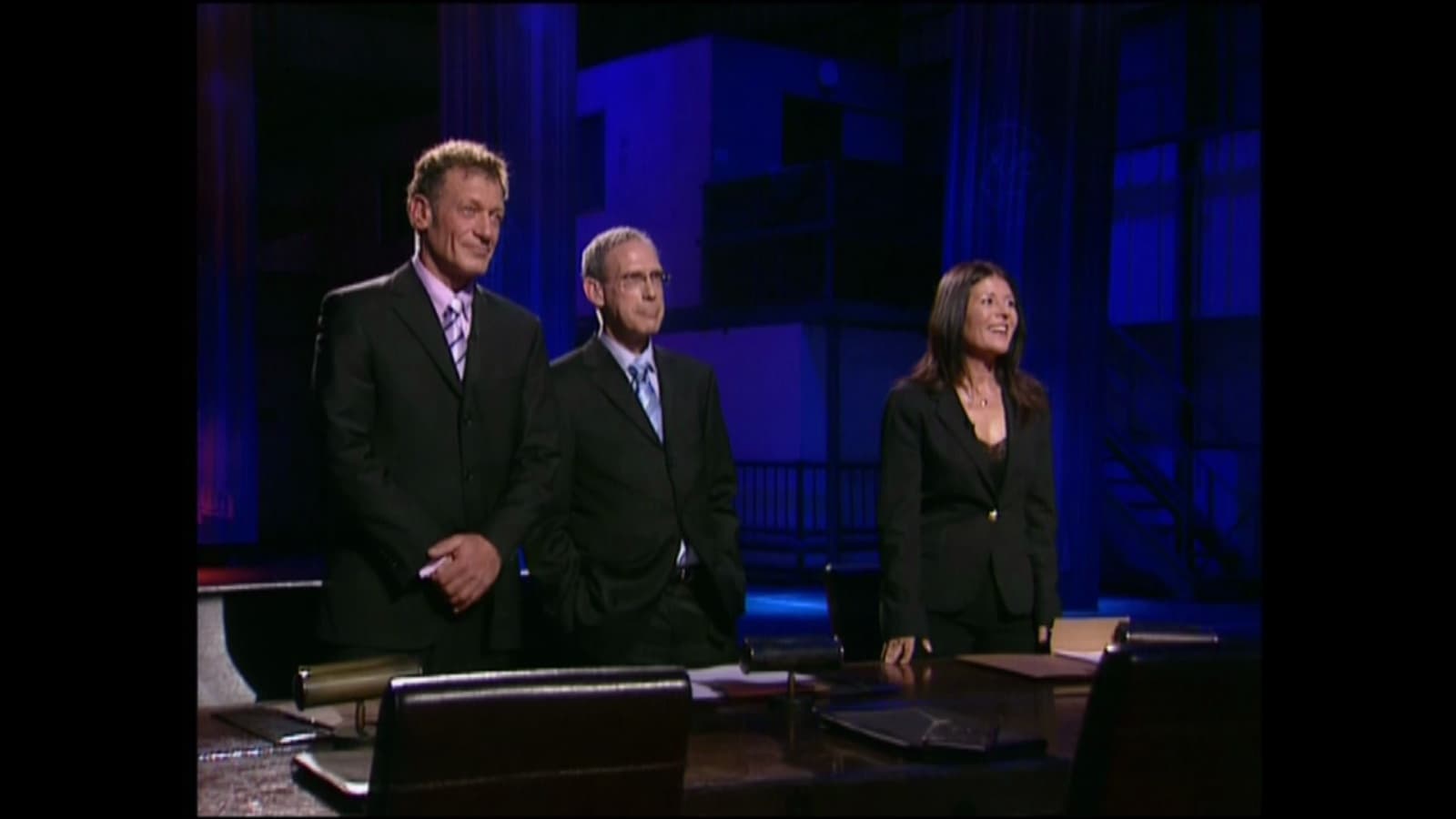 The judges of The Ambassador standing on the blue-lit set.